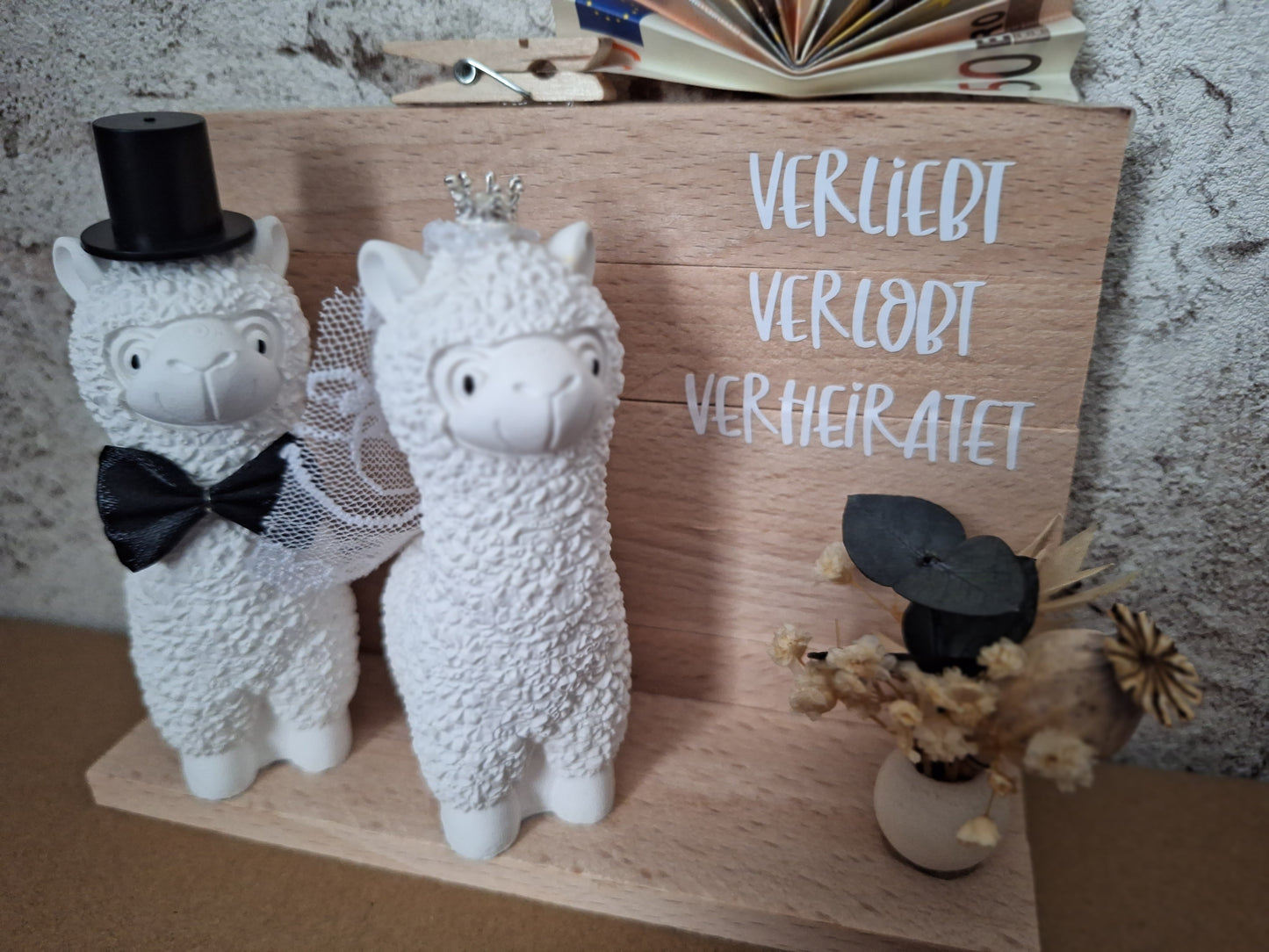 Handgefertigtes Alpaka-Brautpaar als Hochzeitsgeschenk, das Braut mit Krone und Schleier sowie Bräutigam mit Zylinder und Fliege zeigt – liebevolle, personalisierte Dekoration für das Hochzeitsfest. Als Aufschrift steht verliebt, verlobt, verheiratet auf dem Holzaufsteller rechts und darunter eine keine offene Holzkugel mit einem kleinen Trockenblumen darin. Oben Befindet sich eine kleine Klammer für das Geldgeschenk