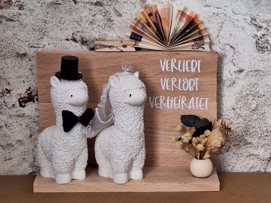 Handgefertigtes Alpaka-Brautpaar als Hochzeitsgeschenk, das Braut mit Krone und Schleier sowie Bräutigam mit Zylinder und Fliege zeigt – liebevolle, personalisierte Dekoration für das Hochzeitsfest. Als Aufschrift steht verliebt, verlobt, verheiratet auf dem Holzaufsteller rechts und darunter eine keine offene Holzkugel mit einem kleinen Trockenblumen darin. Oben Befindet sich eine kleine Klammer für das Geldgeschenk