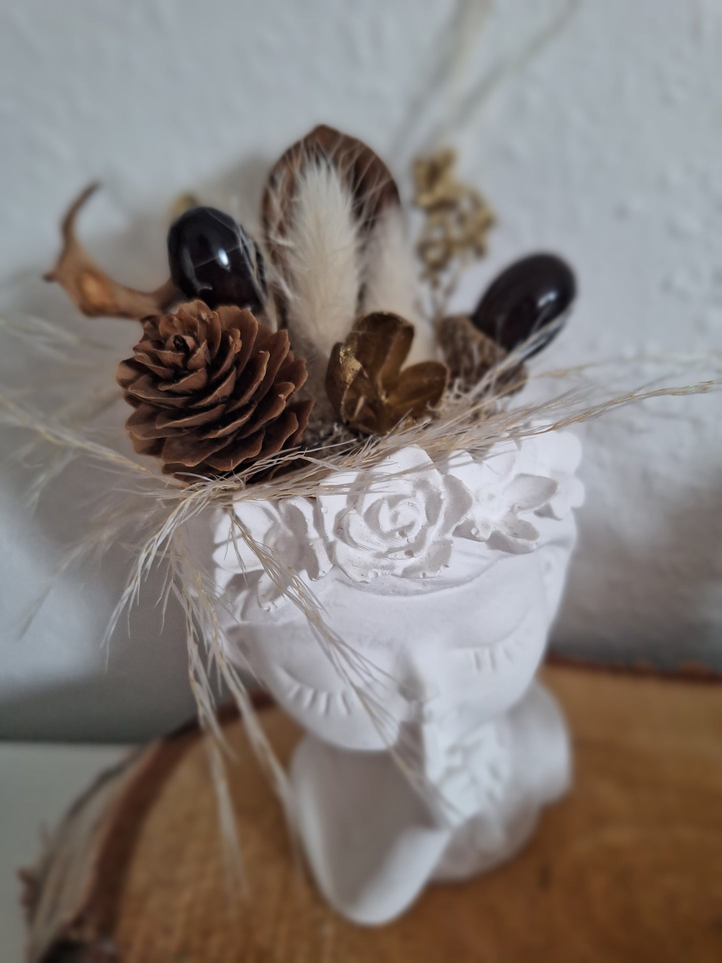 Gezeigt ist ein aus Keraflott gegossener weißer Mädchenkopf ähnlich einer Büste. Das Mädchen zeigt mit einem Finger an den Mund und hat die Augen geschlossen. Am Kopf hat sie ein Blumenrand der als Abschluss dient. Oben auf dem Kopf nach den Blumen stecken Trockenblumen und Kunstdeko im Kopf. Die Farben und Formen sind im Herbstthema braun beige gehalten.