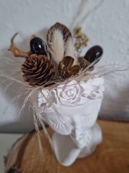 Gezeigt ist ein aus Keraflott gegossener weißer Mädchenkopf ähnlich einer Büste. Das Mädchen zeigt mit einem Finger an den Mund und hat die Augen geschlossen. Am Kopf hat sie ein Blumenrand der als Abschluss dient. Oben auf dem Kopf nach den Blumen stecken Trockenblumen und Kunstdeko im Kopf. Die Farben und Formen sind im Herbstthema braun beige gehalten.