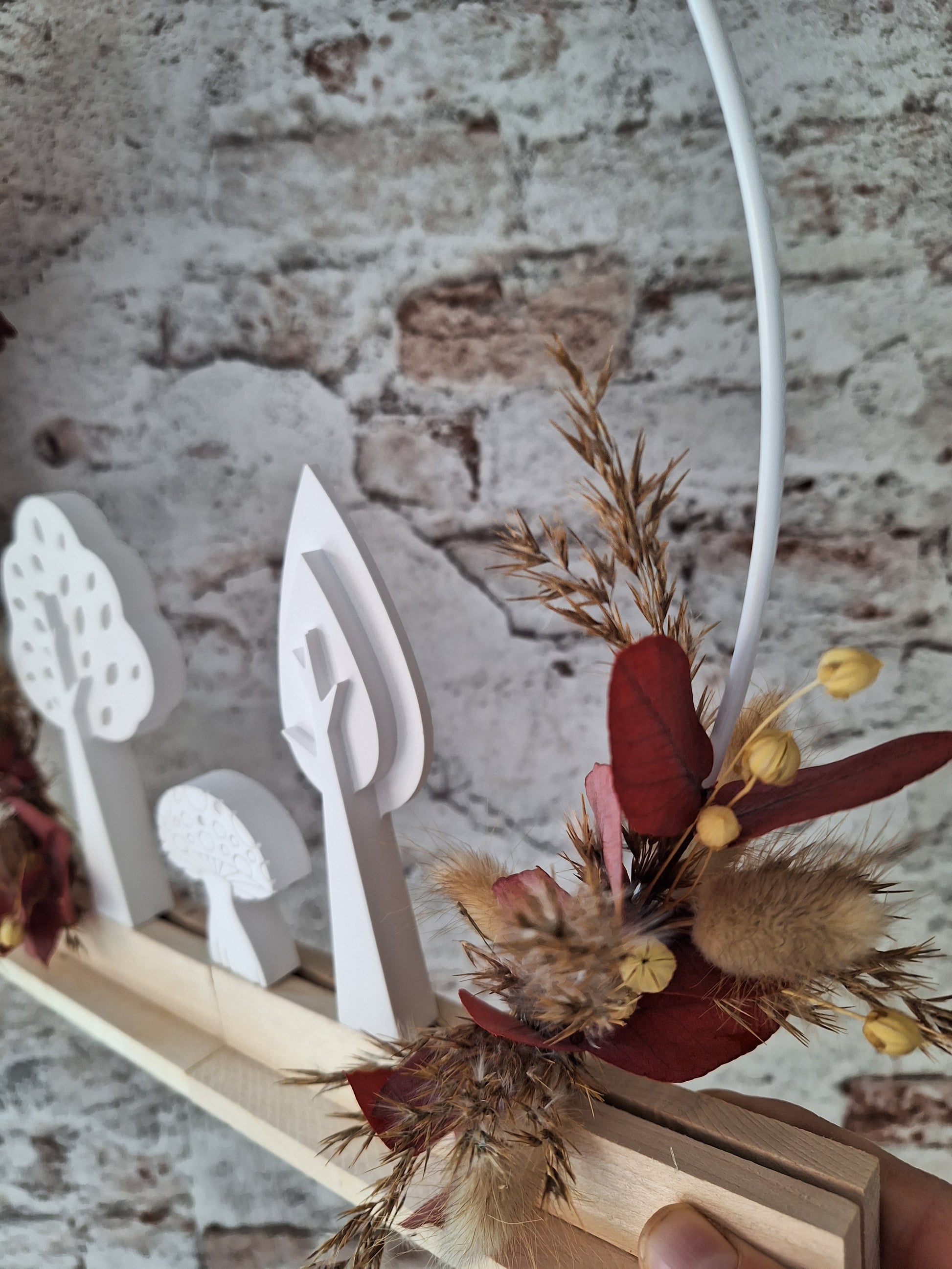 Herbstlicher Dekoaufsteller mit Trockenblumen in Rot- und Brauntönen, weißen Keraflottfiguren in Form von zwei Bäumen und einem Pilz, auf einem Holzsockel helles Holz und Weißer Metallring, handgefertigt und ideal für die saisonale Dekoration.
das bild zeigt eine Nahaufnahme der Trockenblumen und der Figuren von rechts.
