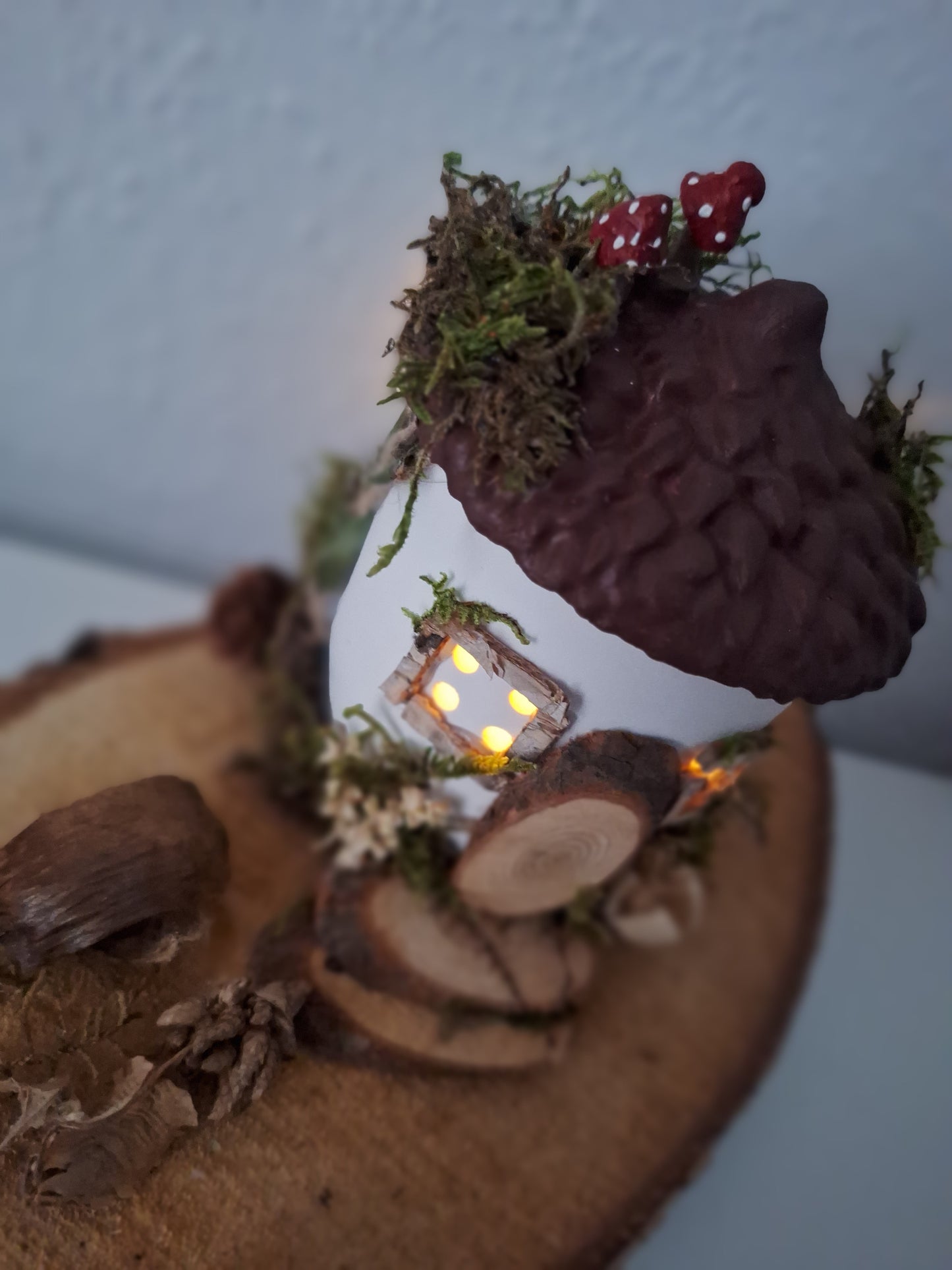 Abgebildet ist ein Feenhäusschen. Es hat vier Fenster mit je vier Löchern und Licht durchzulassen. Die Grundform stellt eine Eichel dar. Der Deckel ist braun. Geschmückt mit getrockneten Moos und Naturmaterialien und zwei handgemachten rot-weißen Pilzen auf dem Dach. Imitierte Tür und Treppe mit Naturmaterialien und Astscheiben. Von inenn leuchtet Licht duch die Fenster.