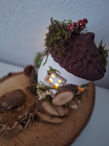 Abgebildet ist ein Feenhäusschen. Es hat vier Fenster mit je vier Löchern und Licht durchzulassen. Die Grundform stellt eine Eichel dar. Der Deckel ist braun. Geschmückt mit getrockneten Moos und Naturmaterialien und zwei handgemachten rot-weißen Pilzen auf dem Dach. Imitierte Tür und Treppe mit Naturmaterialien und Astscheiben. Von inenn leuchtet Licht duch die Fenster.