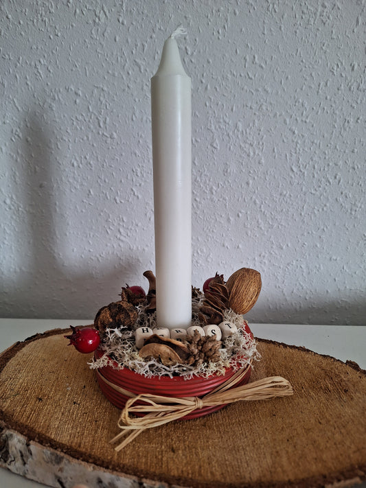 Gezeigt wird ein runder Kerzenständer in rostroter Farbe außen. Um den Rand ist eine Schleife aus Naturmaterialien. Im Innenrand sind durch Steckmasse verschiedene Naturmaterialien und Kunstblumen wie weißes Kunstmoos und eine Rote Beere angebracht. Die Aufschrift Herbst ist mit Holzperlen zwischen den Naturmaterialien angebracht. In der Mitte steckt eine weiße Stabkerze
