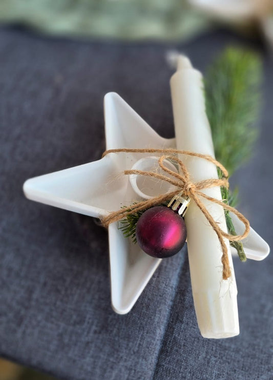 Kerzenhalter in Sternform aus Keraflott. Die Farbe ist weiß. Der Kerzenhalter ist dekoriert mit einer Kerze aus Paraffin, Kunstdekozweig und einer Weihnachtskugel aus Kunststoff befestigt mit einem Juteband am Kerzenhalter.