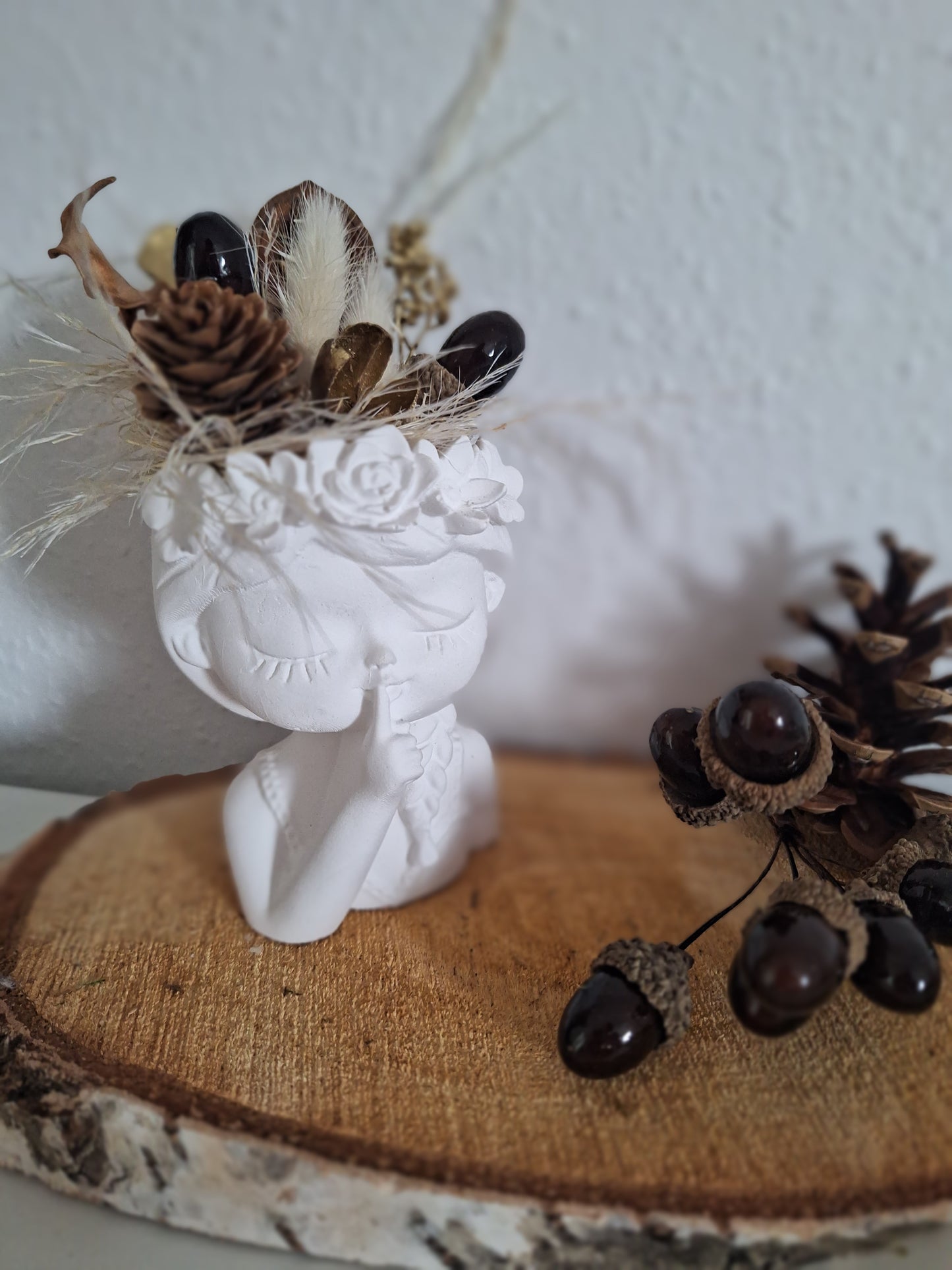 Gezeigt ist ein aus Keraflott gegossener weißer Mädchenkopf ähnlich einer Büste. Das Mädchen zeigt mit einem Finger an den Mund und hat die Augen geschlossen. Am Kopf hat sie ein Blumenrand der als Abschluss dient. Oben auf dem Kopf nach den Blumen stecken Trockenblumen und Kunstdeko im Kopf. Die Farben und Formen sind im Herbstthema braun beige gehalten.