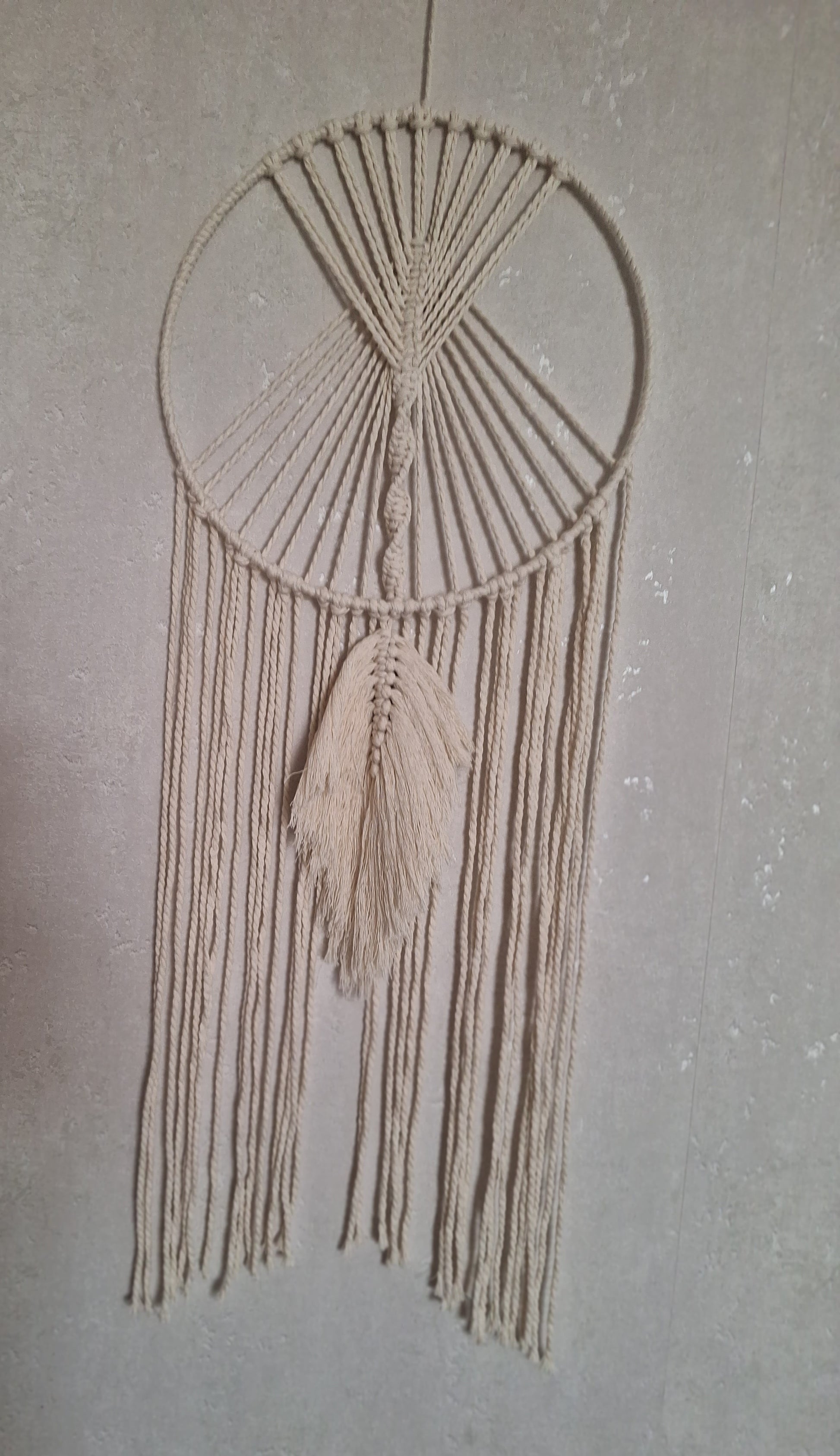 Handgefertigte cremefarbene Makramee-Wanddekoration an einem eleganten Metallring, ideal für Boho- und moderne Wohnräume, bringt Wärme und Stil in dein Zuhause. Die Farbe des Makramee Garns ist beige. der Metallring ist mit Garn umwickelt. Unter dem Ring hängen Makramee Garn Schnüre hinab und mittig eine Makramee Feder in locker fallend