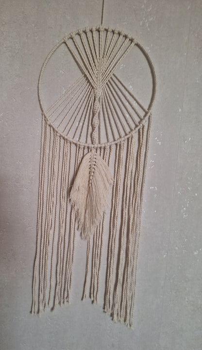 Handgefertigte cremefarbene Makramee-Wanddekoration an einem eleganten Metallring, ideal für Boho- und moderne Wohnräume, bringt Wärme und Stil in dein Zuhause. Die Farbe des Makramee Garns ist beige. der Metallring ist mit Garn umwickelt. Unter dem Ring hängen Makramee Garn Schnüre hinab und mittig eine Makramee Feder in locker fallend