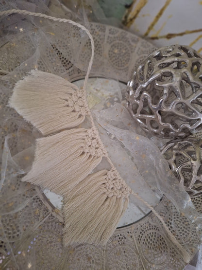 Gezeigt ist eine Makramee Wimpelkette im Boho-Stil in beige. Es sind drei Flechtteile bei diesem Beispiel. oben ist zu einem Dreieck geknüpft und der Rest fällt glatt zu einer leichten Spitze herab.