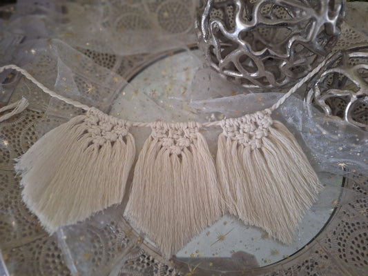 Gezeigt ist eine Makramee Wimpelkette im Boho-Stil in beige. Es sind drei Flechtteile bei diesem Beispiel. oben ist zu einem Dreieck geknüpft und der Rest fällt glatt zu einer leichten Spitze herab.