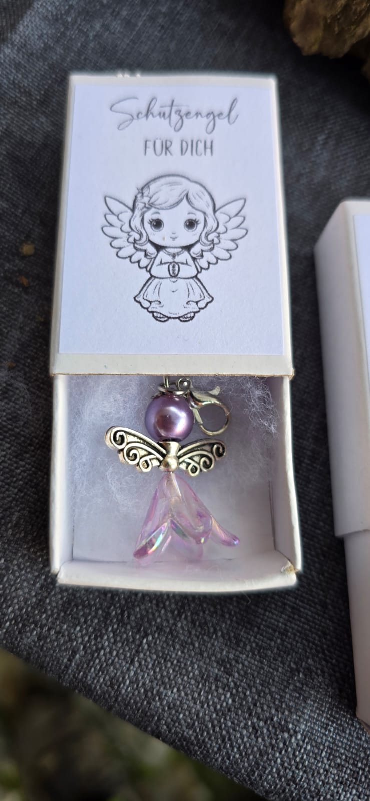 Kleiner Schutzengelanhänger gefertigt aus Perlen. Liegt in einer kleinen Schachtel gebettet auf Watte. Auf der Schachtel ist ein Aufkleber mit dem Spruch "Schutzengel für dich" und einem Bild eines kleinen Engelmädchens.