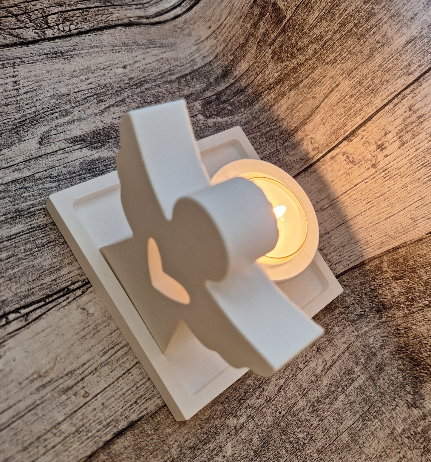 Weißes Keraflott-Set bestehend aus einem gegossenen Engel, einem kleinen viereckigen Tablett und einem Teelichthalter. Das handgefertigte Set lässt durch das brennende Teelicht ein dezentes, elegantes Licht durch das Herz-Loch im Engel und den Hintergrund scheinen, wodurch eine warme und ruhige Atmosphäre entsteht.