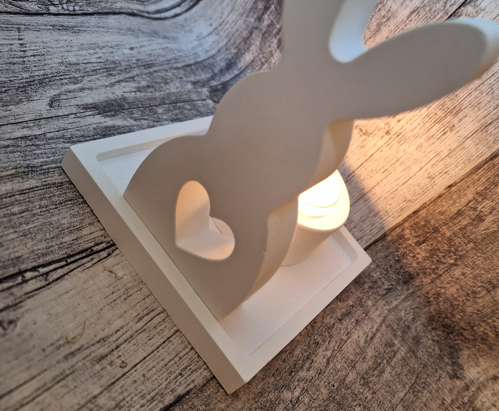 Weißes Keraflott-Set bestehend aus einem gegossenen Hasen, einem kleinen viereckigen Tablett und einem Teelichthalter. Das Set ist handgefertigt und verleiht mit dem Licht des Teelichts ein dezentes, elegantes Leuchten durch das Herz-Loch im Hasen und den Hintergrund.