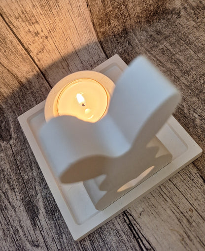 Weißes Keraflott-Set bestehend aus einem gegossenen Hasen, einem kleinen viereckigen Tablett und einem Teelichthalter. Das Set ist handgefertigt und verleiht mit dem Licht des Teelichts ein dezentes, elegantes Leuchten durch das Herz-Loch im Hasen und den Hintergrund.