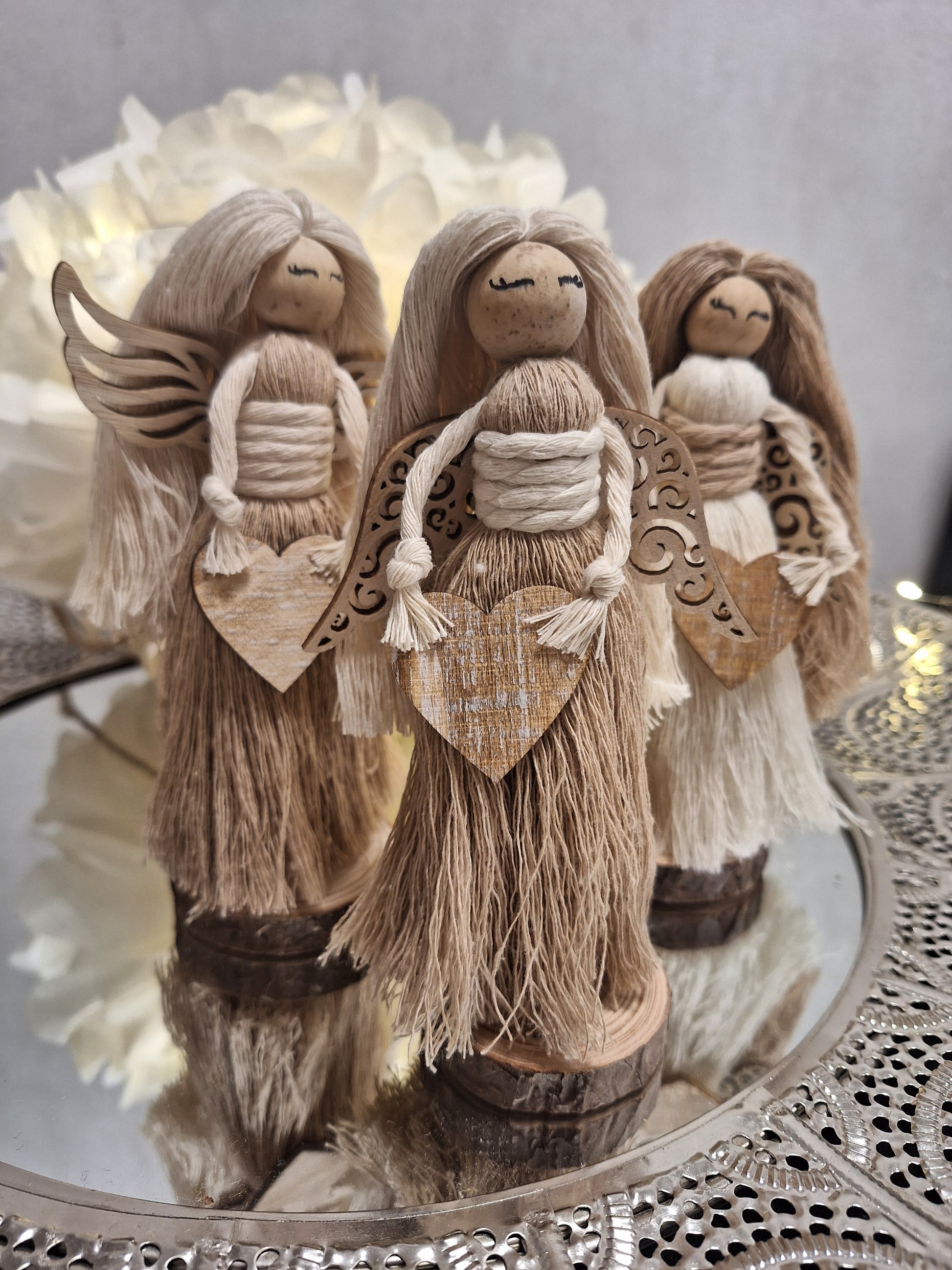 Gezeigt sind drei kleine Engel. Die Engel stehen auf einem Holzständer im Inneren. Man sieht nur einen kleinen Sockel. Die Haare sind glatt und lang aus Makramee-Garn gekämmt. Als Kopf dient eine Holzkugel woran die Haare begestigt sind. Der Oberkörper ist auch aus gekämmten Makramee-Garn befestigt mit einem Gürtel aus Makramee am Oberkörper. Die Arme sind aus Makramee. Die Hände sind durch Knoten angedeutet und halten ein Herz aus Holz. Die Farben sind beige/hellbraun; beige/hellbr.; braun/beige